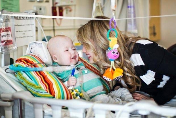 Implante respiratório criado em impressora 3D cura bebe. Mãe de bebe sorrindo para ele em quarto de hospital. Bebê sorrindo para mãe com roupinha listrada de branco e verde água.