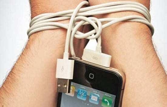 Mãos humanas amarradas com cabo de carregador do Iphone e smartphone pendurado entre os braços da pessoa.